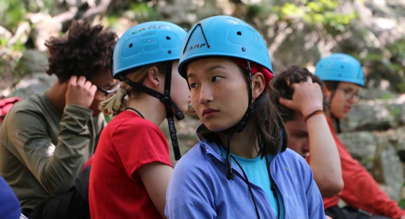 A group of young people appear to prepare to rock climb. A few of them are wearing helmets. 